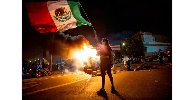 Una mexicana ondea su bandera mientras las llamas salen de un contenedor de basura durante una de las protestas en Los Ángeles, California, contra las redadas y la militarización ordenadas por Donald Trump. Sábado 8 de junio de 2025. Foto: AP/Ethan Swope